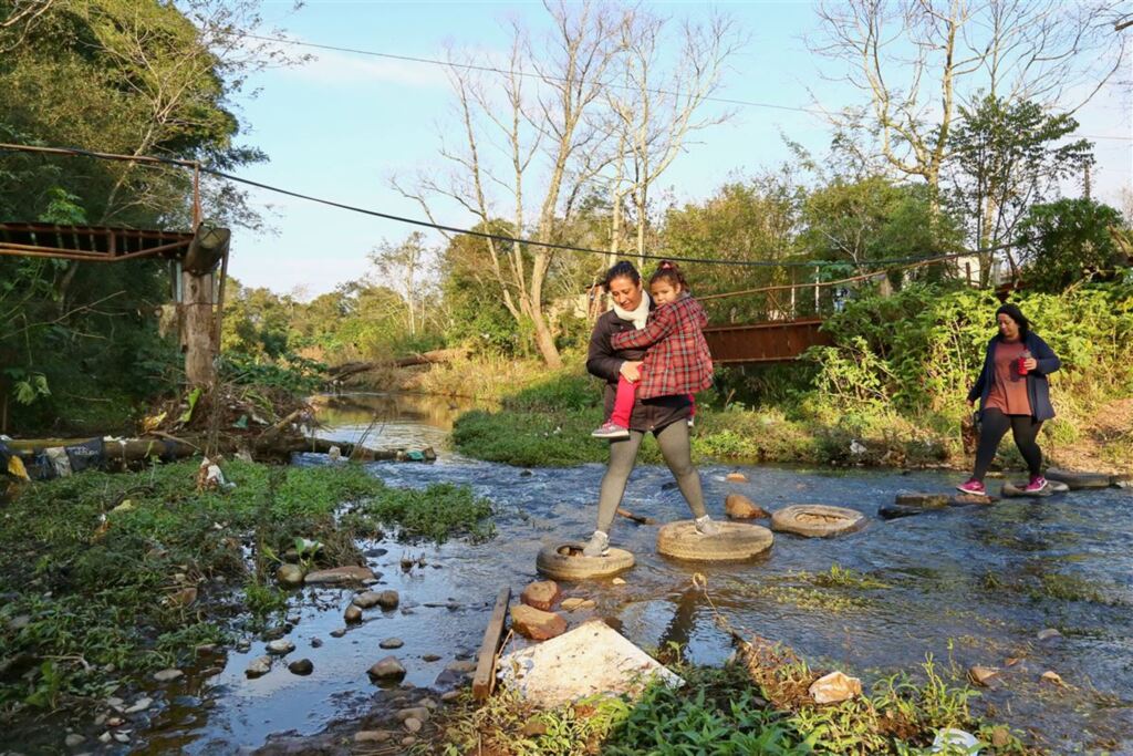 Foto: Charles Guerra (Diário) - Patrícia da Rosa, com a filha Laura, 5 anos, no colo e a irmã Simone da Rosa cruzam o rio sobre uma passarela feita de pneus e pedras
